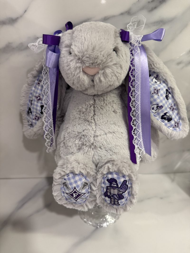 A grey plush bunny on a glass stand, with purple and lace ribbons tied to each ear and checked fabric on the inner ears and paw pads.